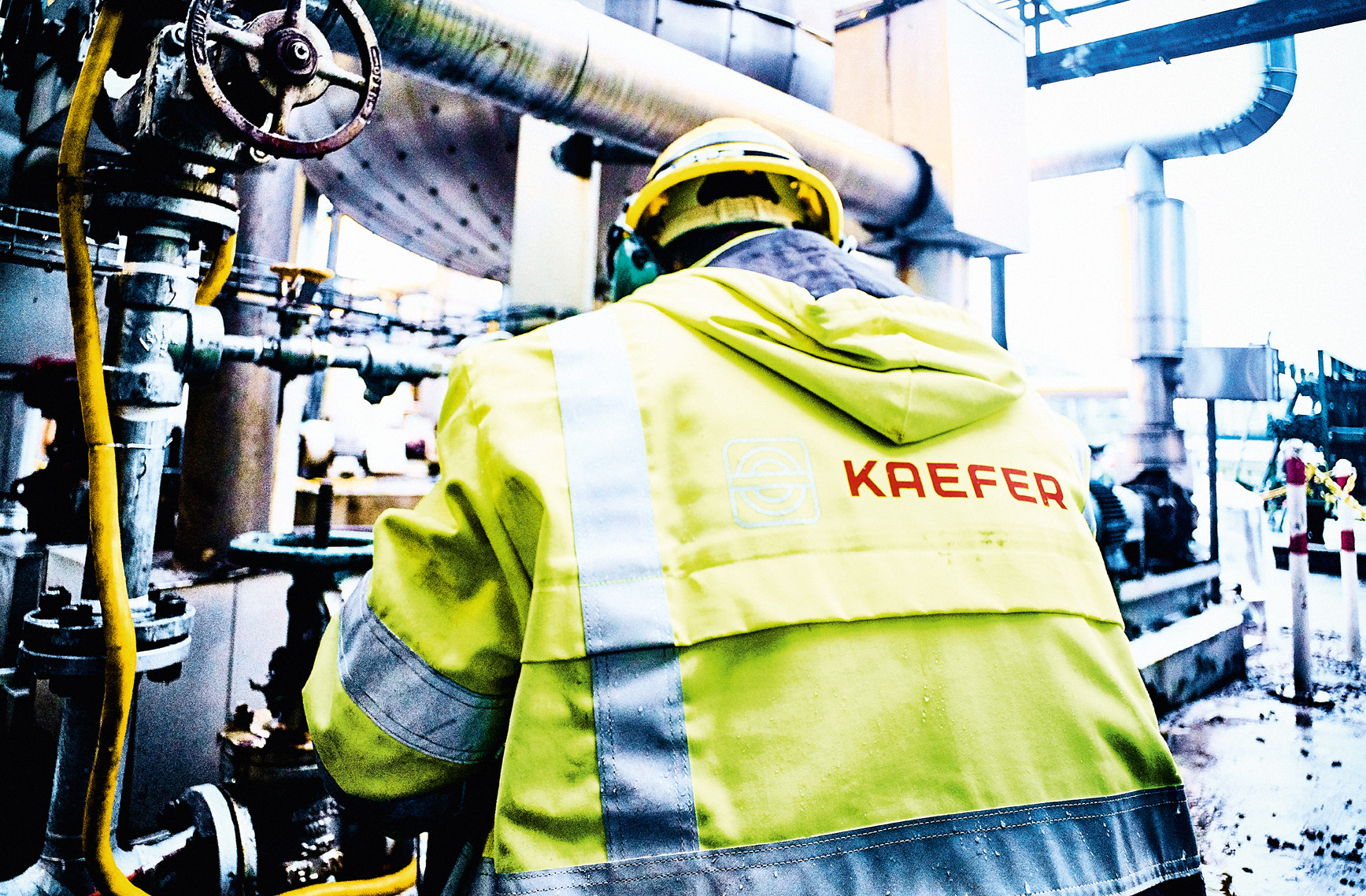 Rückansicht eines KAEFER-Mitarbeiters in neongelber Warnschutzkleidung mit Firmenlogo, der Wartungsarbeiten an einem komplexen Rohrleitungssystem mit Absperrventilen durchführt