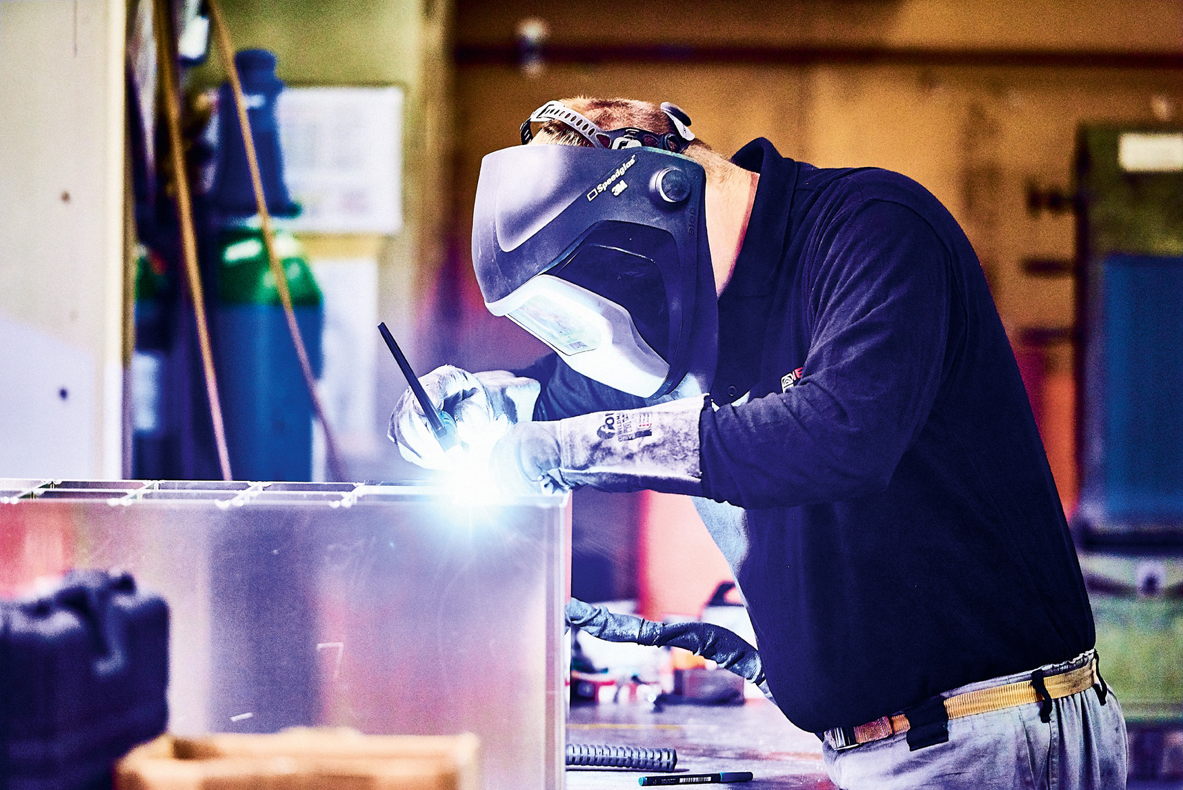 Ein Metallbauer mit Schutzausrüstung und Automatik-Schweißhelm führt präzise Schweißarbeiten an einem Edelstahlgehäuse durch, während der Lichtbogen die Arbeitsstelle hell erleuchtet