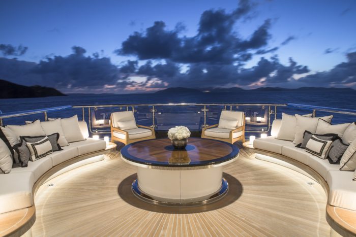 Beleuchteter Loungebereich mit halbkreisförmigen Sofas und einem runden Holztisch auf einem Schiffsdeck bei Abenddämmerung