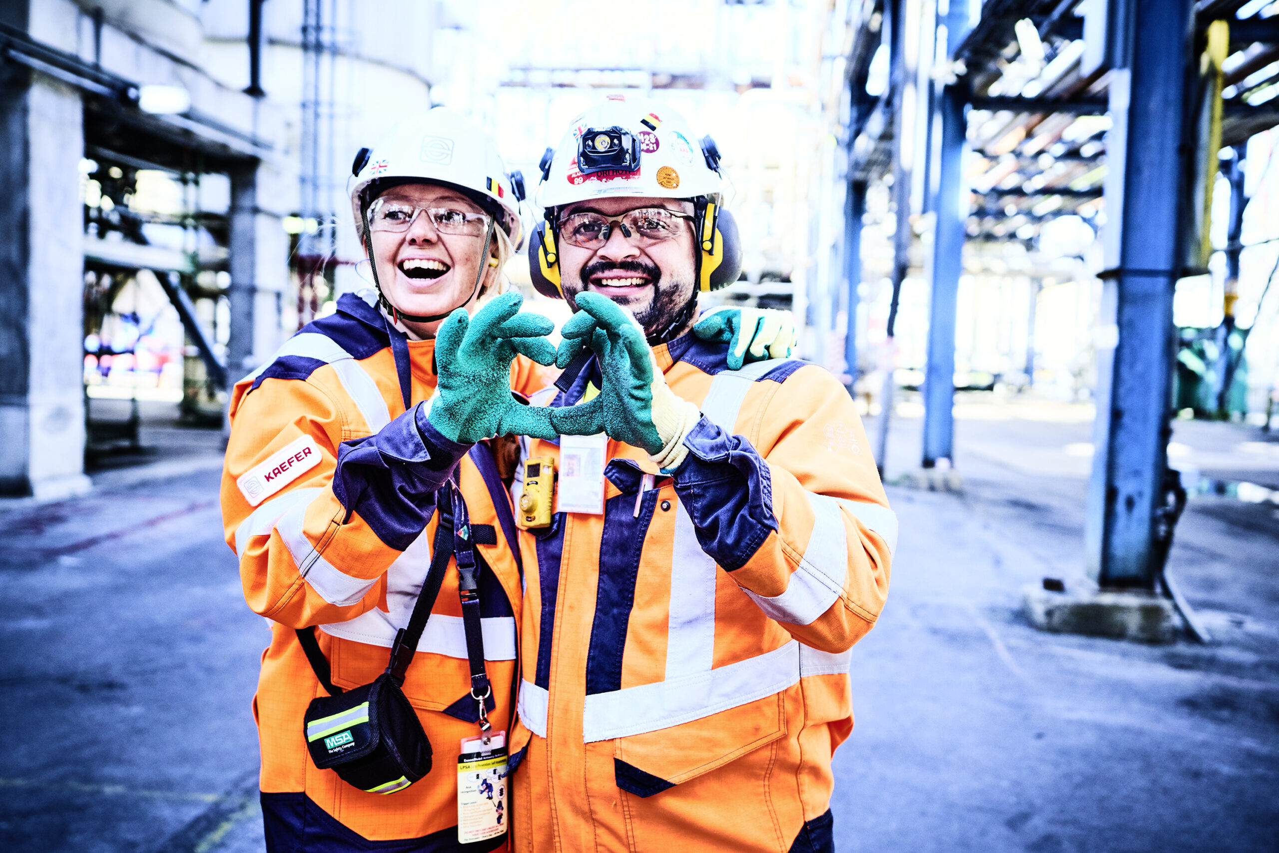 Eine lachende KAEFER Mitarbeiterin und ein Kollege in orangefarbener Warnschutzkleidung und Schutzhelmen formen gemeinsam mit ihren behandschuhten Händen ein Herz-Symbol vor einer Industrie-Kulisse