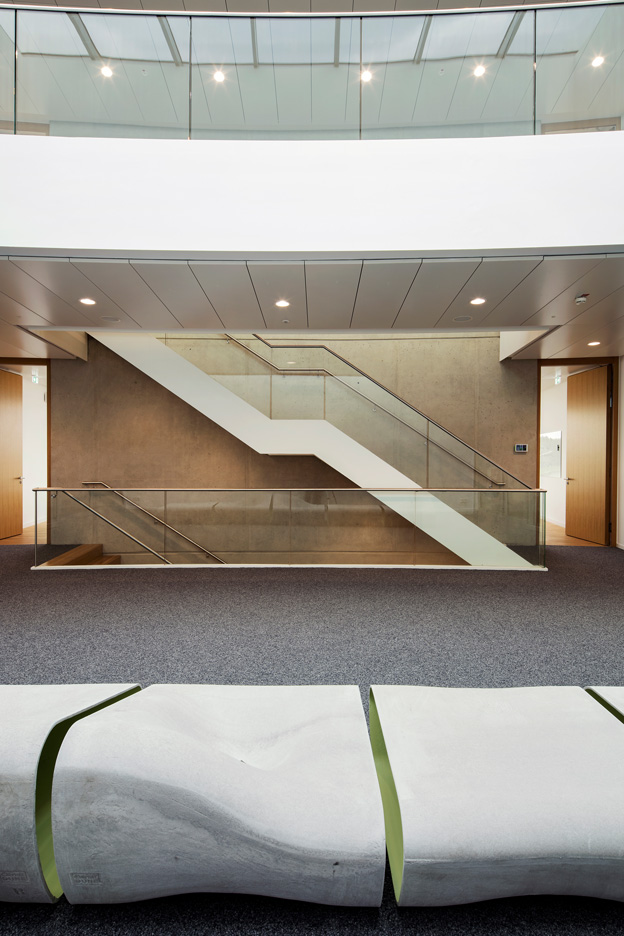 Blick auf eine minimalistische, weiße Treppenkonstruktion mit Glasgeländer vor einer Sichtbetonwand in einem Bürogebäude