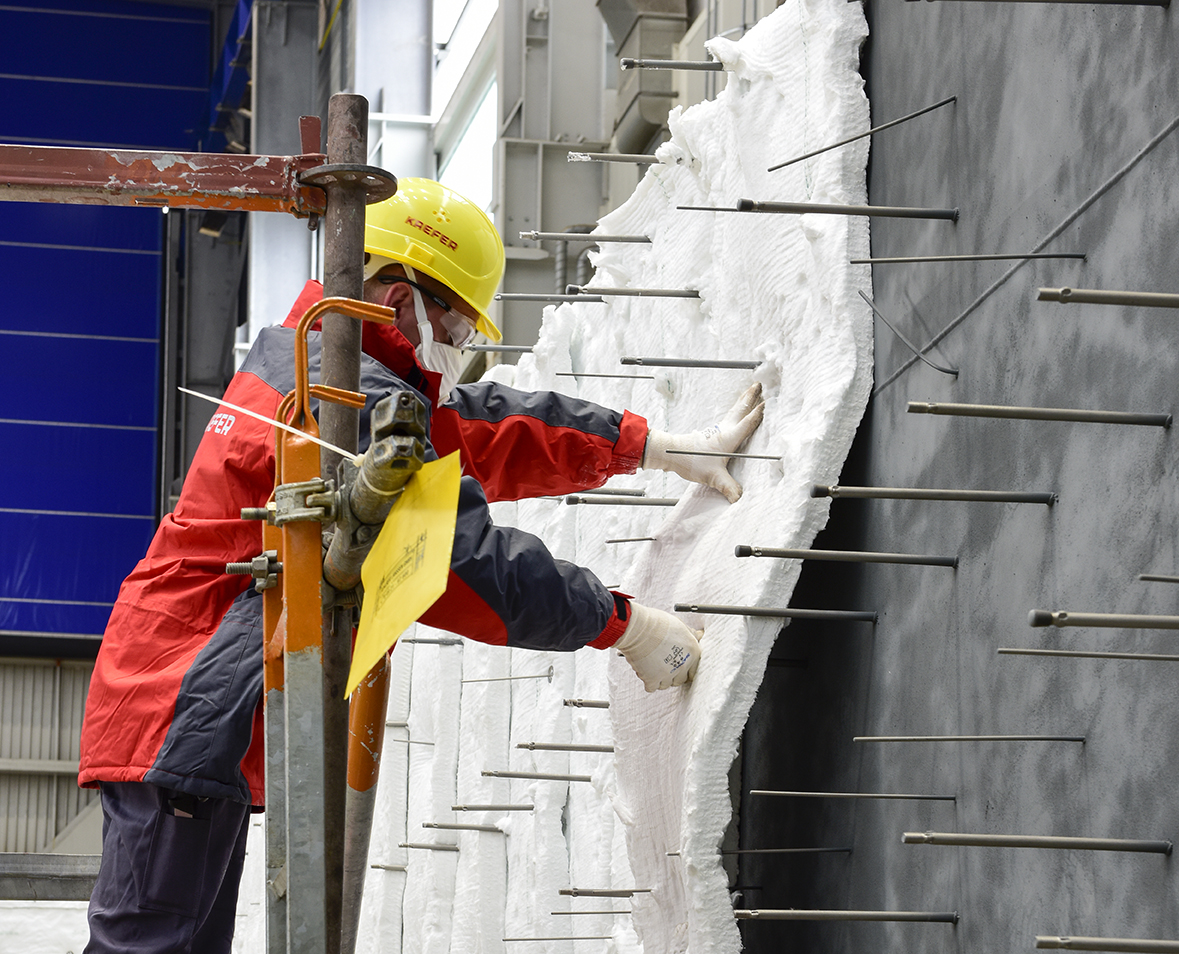 Ein Arbeiter mit Atemschutzmaske und Schutzbrille bringt Dämmmatten an einer Wand an, die durch zahlreiche horizontal herausstehende Metallstifte fixiert werden