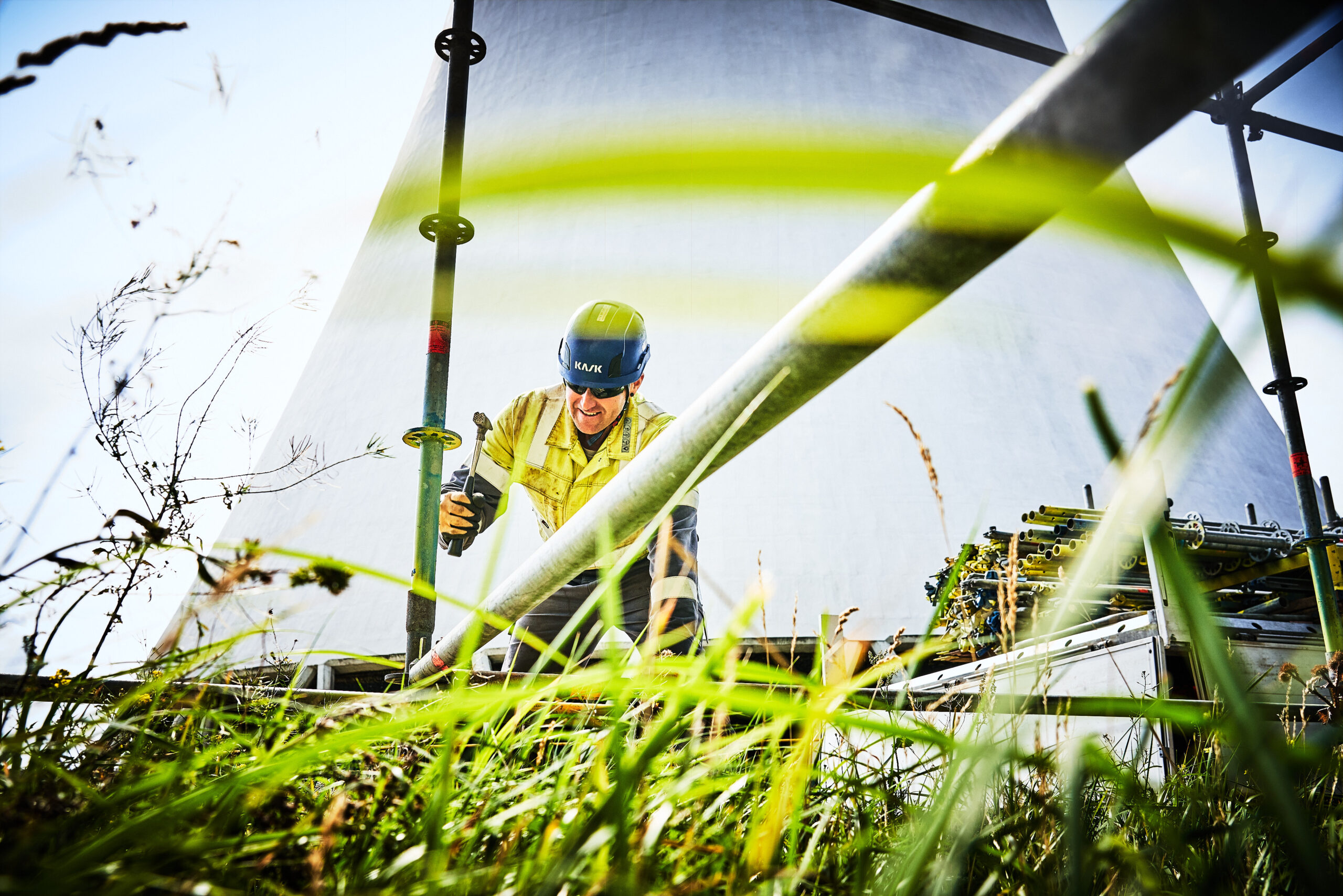Froschperspektive durch Grashalme auf einen KAEFER Mitarbeiter in Arbeitskleidung, welcher ein Gerüst aufbaut