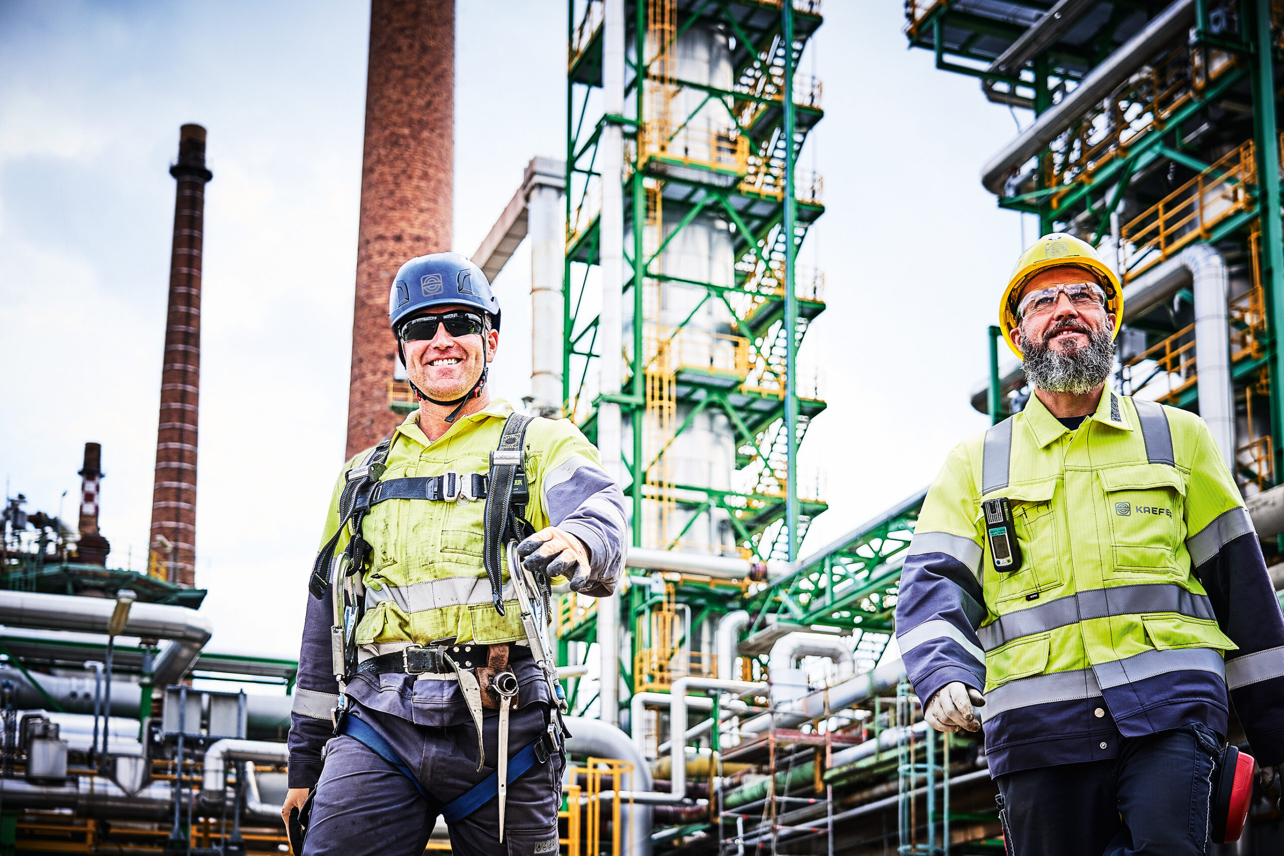 Zwei lächelnde KAEFER Mitarbeiter in Arbeitsanzügen auf einer Industrieanlage