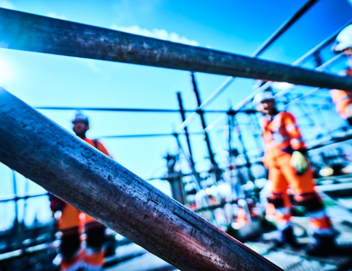 Verschwommene Sicht durch Gerüststangen auf drei KAEFER Arbeiter in Arbeitsanzügen