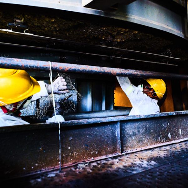 Zwei Mitarbeiter in weißer Schutzkleidung und gelben Helmen befestigen Drahtgeflecht an einer Industrie-Isolierung