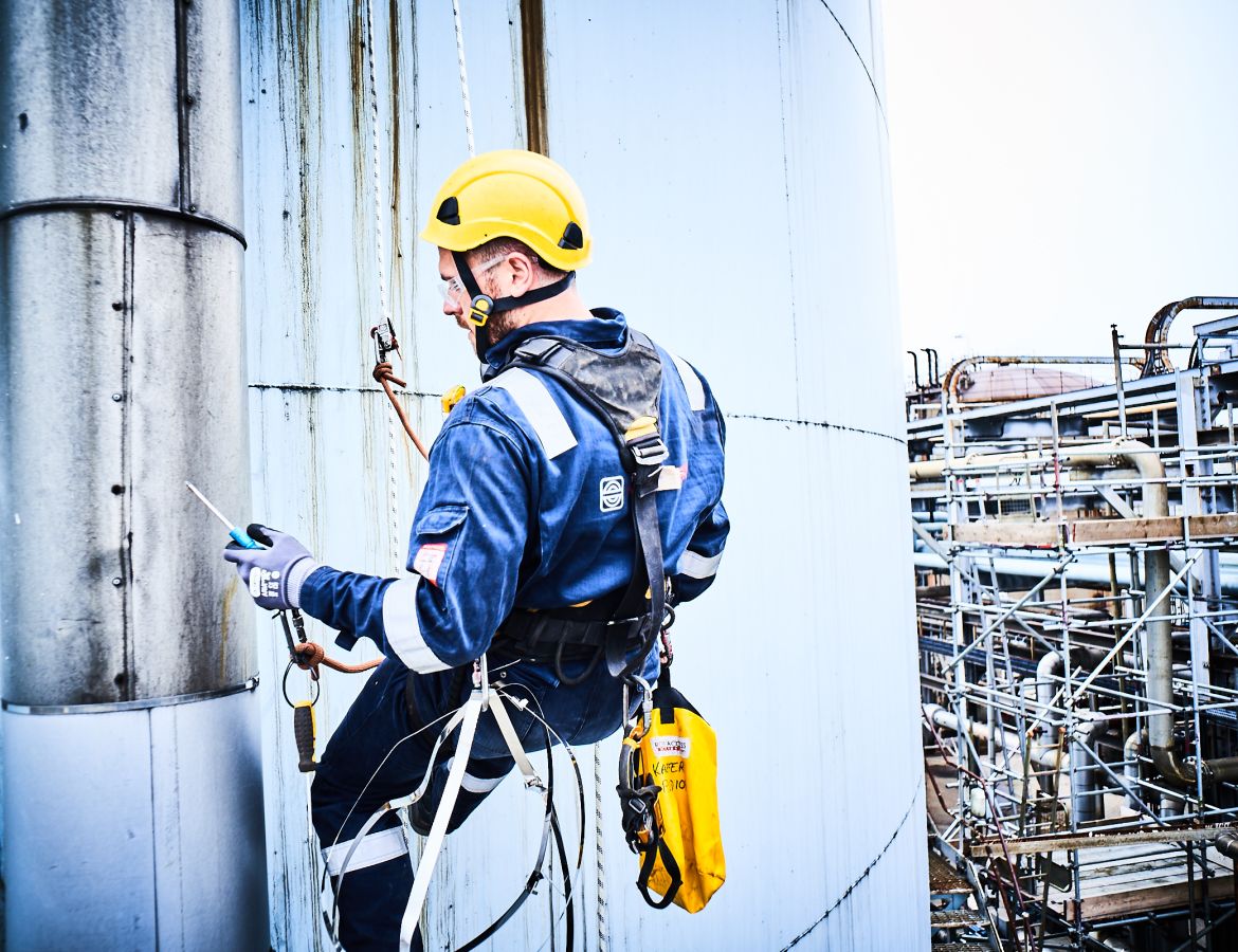 Ein Industriekletterer mit Schutzhelm hängt in den Seilen und führ tArbeiten an der Außenwand eines großen Silos durch