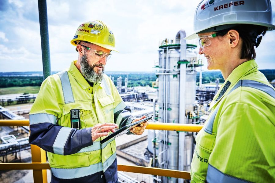 Zwei KAEFER-Mitarbeiter in Schutzausrüstung besprechen Arbeitsdaten auf einem Tablet vor der Kulisse einer Industrieanlage