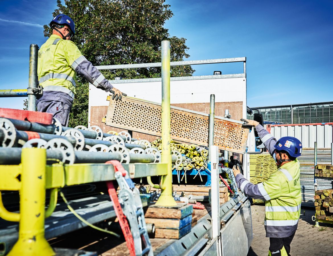 Ein KAEFER Mitarbeiter in Arbeitskleidung reicht einem anderem Arbeiter ein Gerüstteil von einem Transporter herunter
