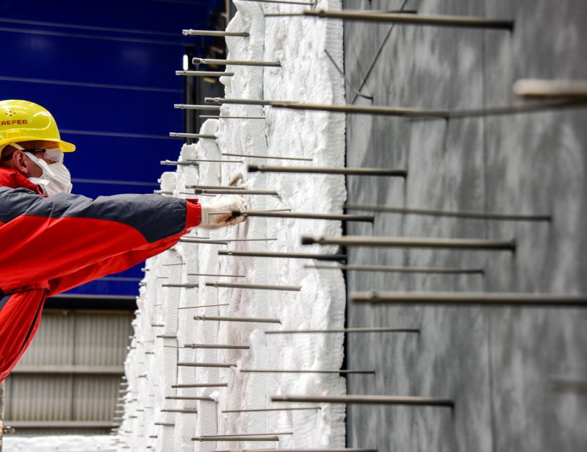 Ein Arbeiter mit Atemschutzmaske und Schutzbrille bringt Dämmmatten an einer Wand an, die durch zahlreiche horizontal herausstehende Metallstifte fixiert werden