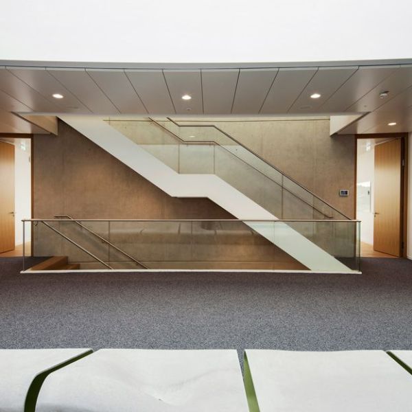 Blick auf eine minimalistische, weiße Treppenkonstruktion mit Glasgeländer vor einer Sichtbetonwand in einem Bürogebäude