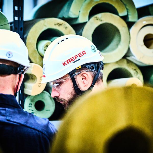 Zwei KAEFER-Mitarbeiter mit Schutzhelmen vor einem Lagerregal mit runden Dämmstoff-Formteilen