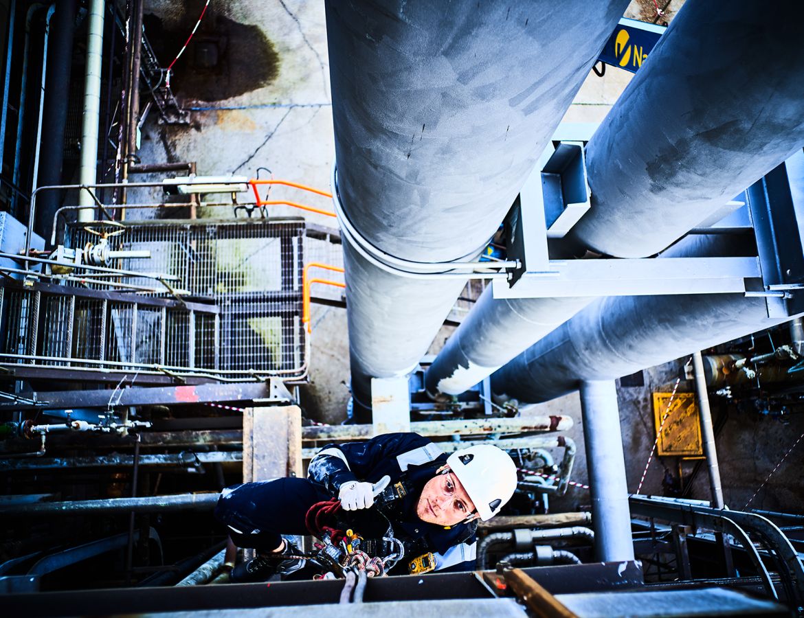 Perspektive von oben auf einen Industriekletterer, der an Seilen gesichert über einer Industrieanlage mit großen Rohrleitungen arbeitet