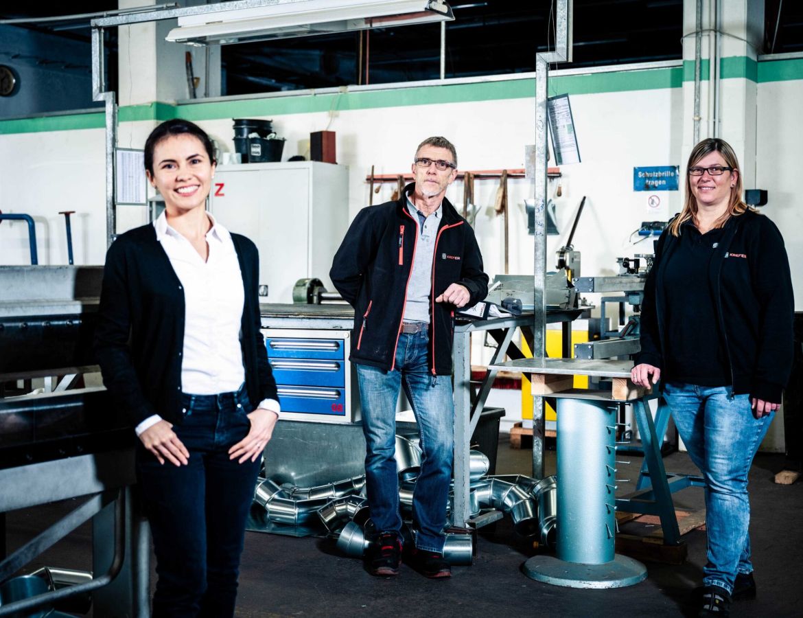 Drei KAEFER Mitarbeiter der Industrieisolierung posieren lächelnd in einer Fertigungshalle vor Werkbänken und Blechbauteilen