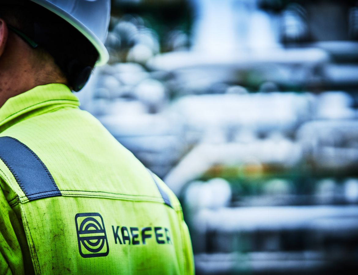 Nahaufnahme des Rückens eines KAEFER Mitarbeiters, wo das Logo auf der gelben Arbeitskleidung zu sehen ist. Im Hintergrund ist eine verschwommene Industrielandschaft