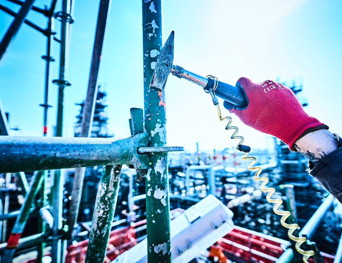 Nahaufnahme einer Hand in einem roten Schutzhandschuh, die mit einem Hammer einen Keil an einem Gerüstpfosten festschlägt