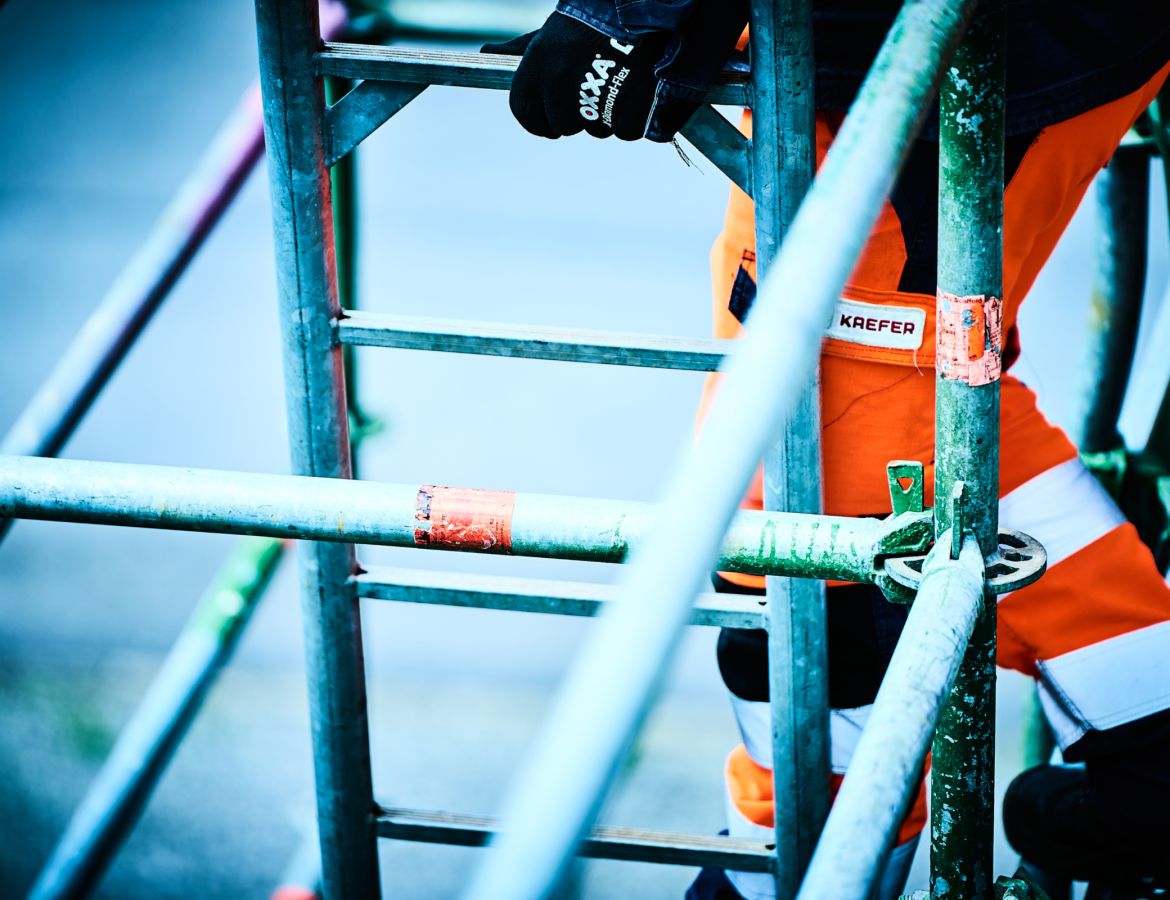 Detailaufnahme eines KAEFER Mitarbeiters in Arbeitskleidung, der eine Metallleiter an einem Gerüst emporsteigt